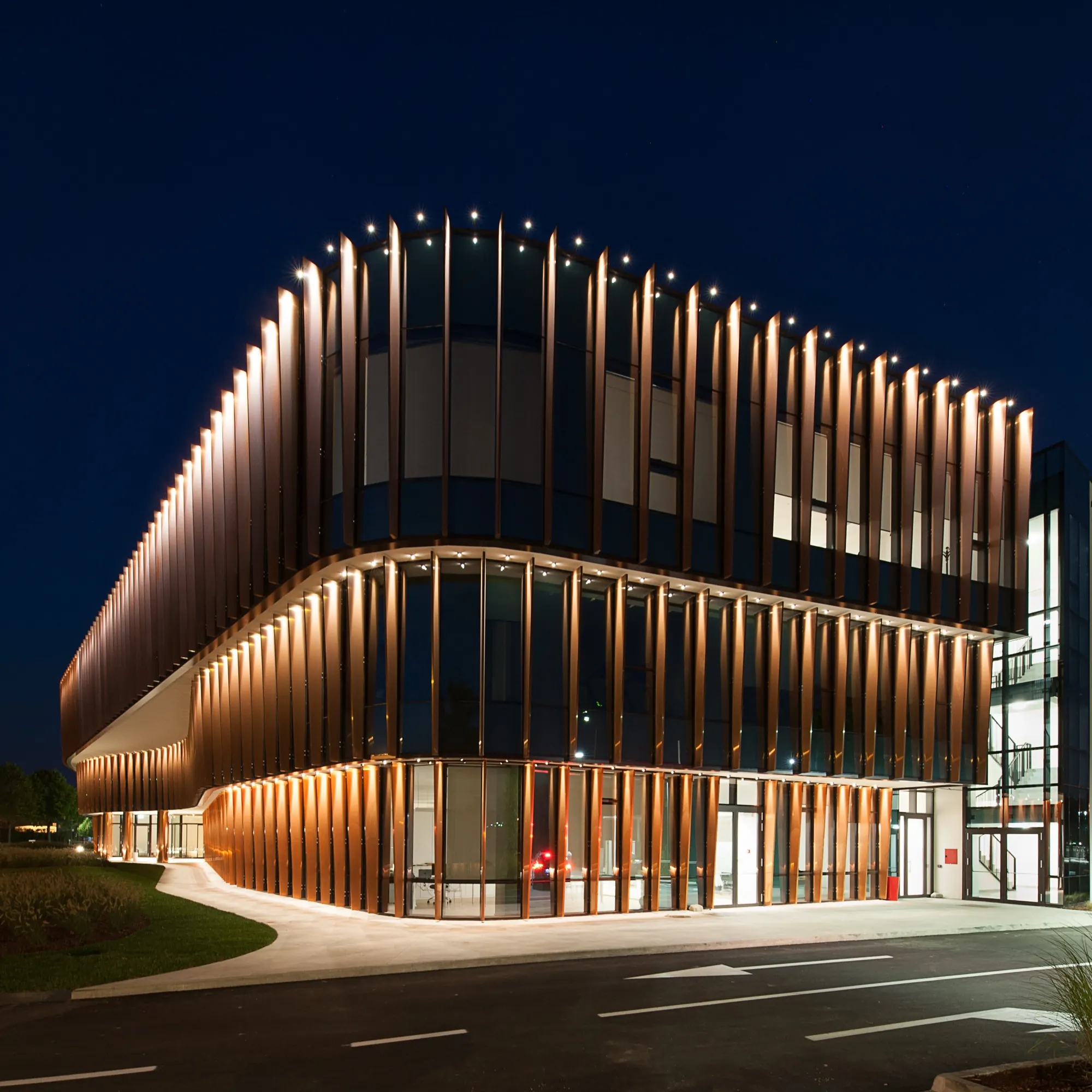 Corner of a two-storey building wrapped in warm-lit vertical fins, lit glazing behind