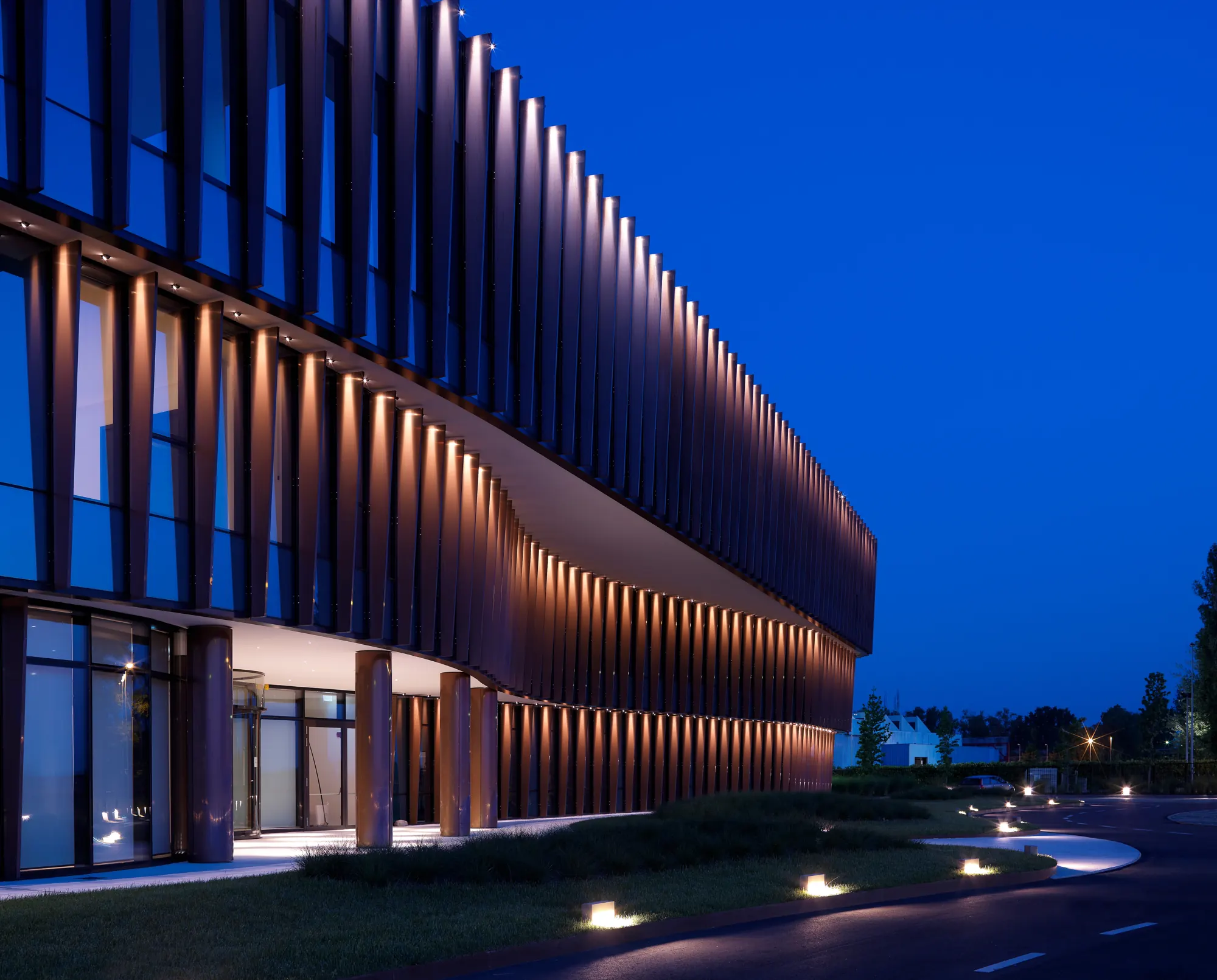 A curved facade of lit copper fins at night, recessed ground lights tracing the lawn edge