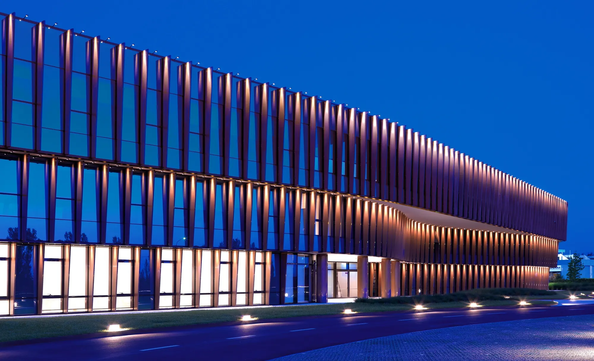 Copper vertical fins on a long office facade at blue hour, each fin grazed from its base, in-ground lights along the drive