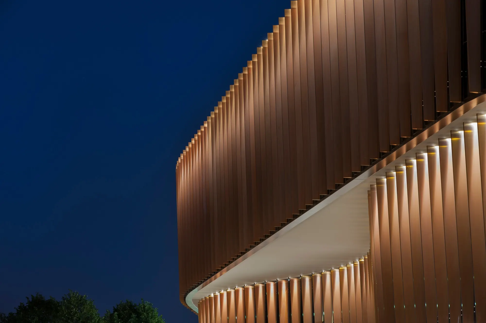 The underside of a cantilevered fin facade, warm light rising along each fin into the dusk