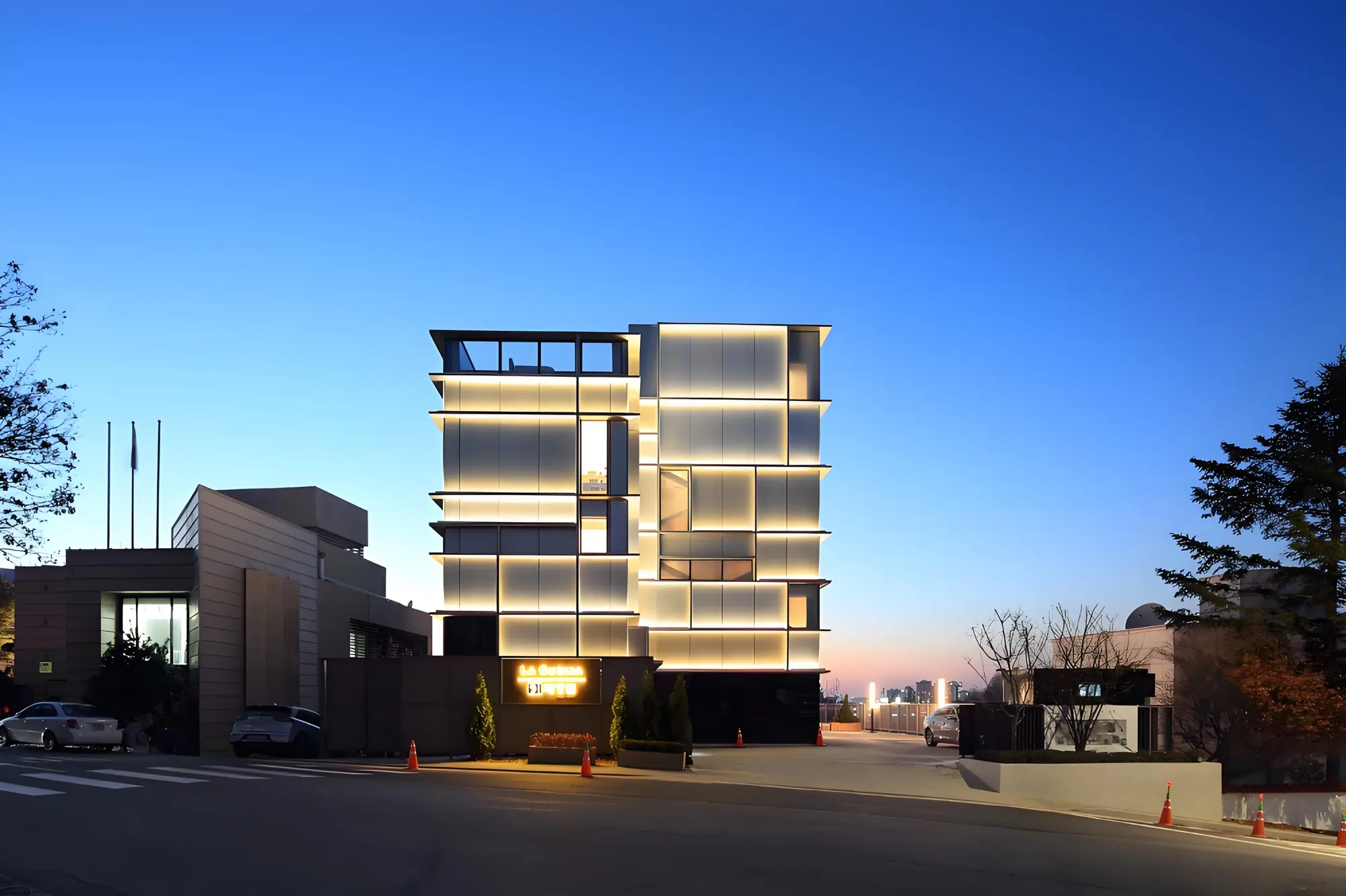 A small glass building on a street corner at dusk, each floor band lit with warm linear light